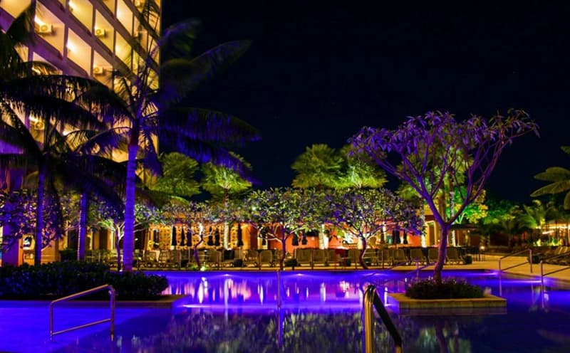Piscina do resort à noite com o prédio e coqueiros iluminados.