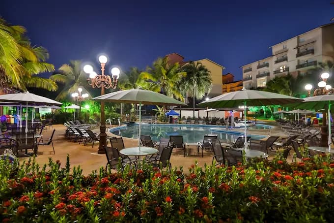Área da piscina com mesas, cadeiras e iluminação noturna.