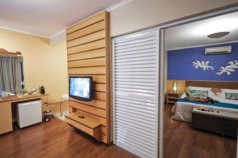 Quarto de hotel moderno com TV, frigobar e porta para o banheiro.