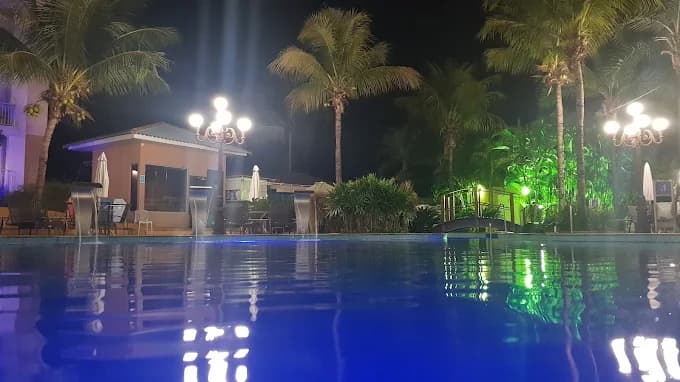 Borda da piscina iluminada à noite, com coqueiros ao redor.