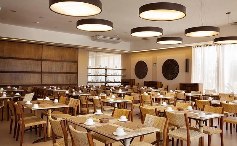 Amplo salão do restaurante do hotel com mesas redondas e luminárias circulares.