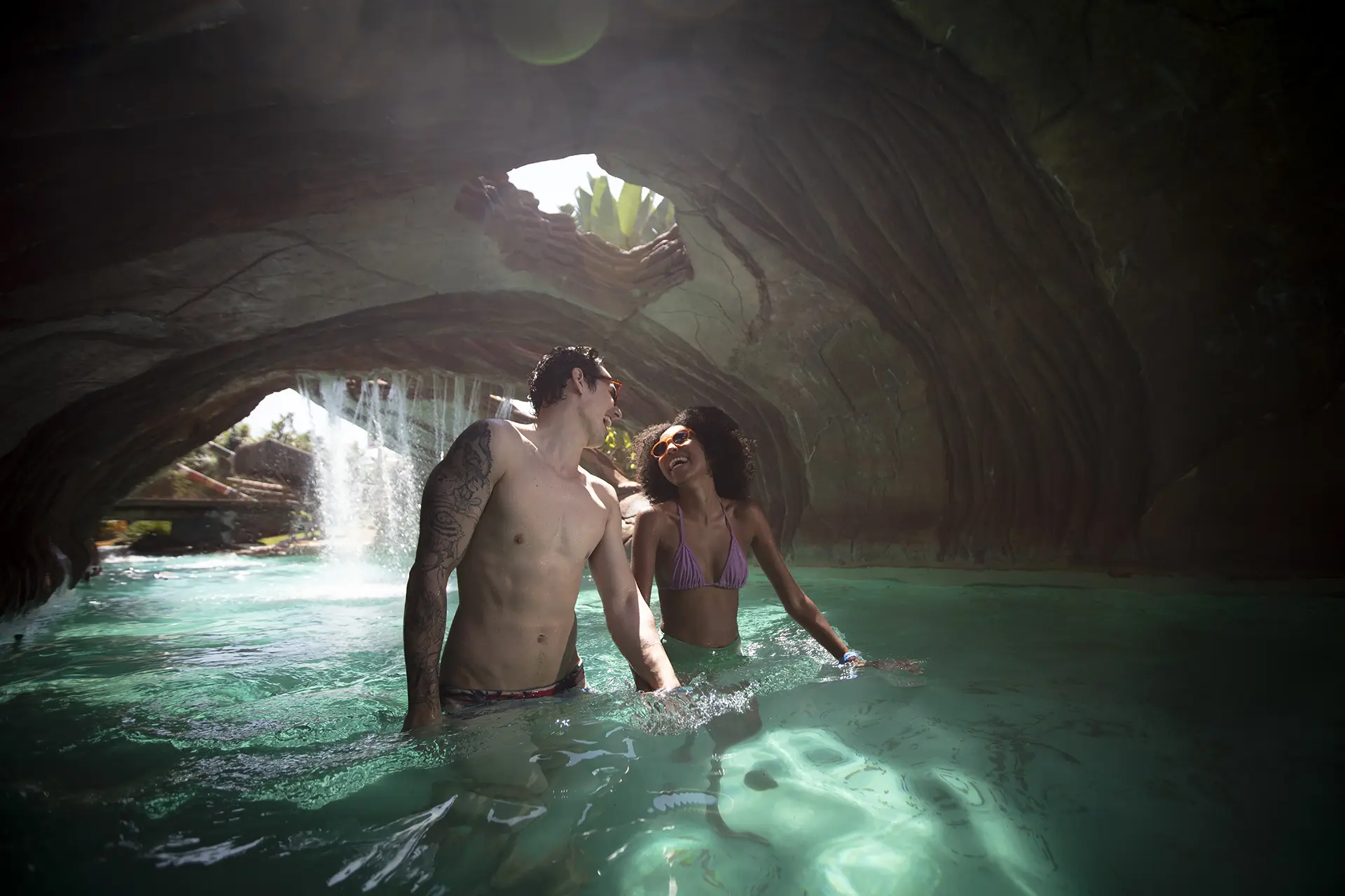 Pessoas conversando e relaxando na borda de uma piscina com paisagismo de pedras e coqueiros.