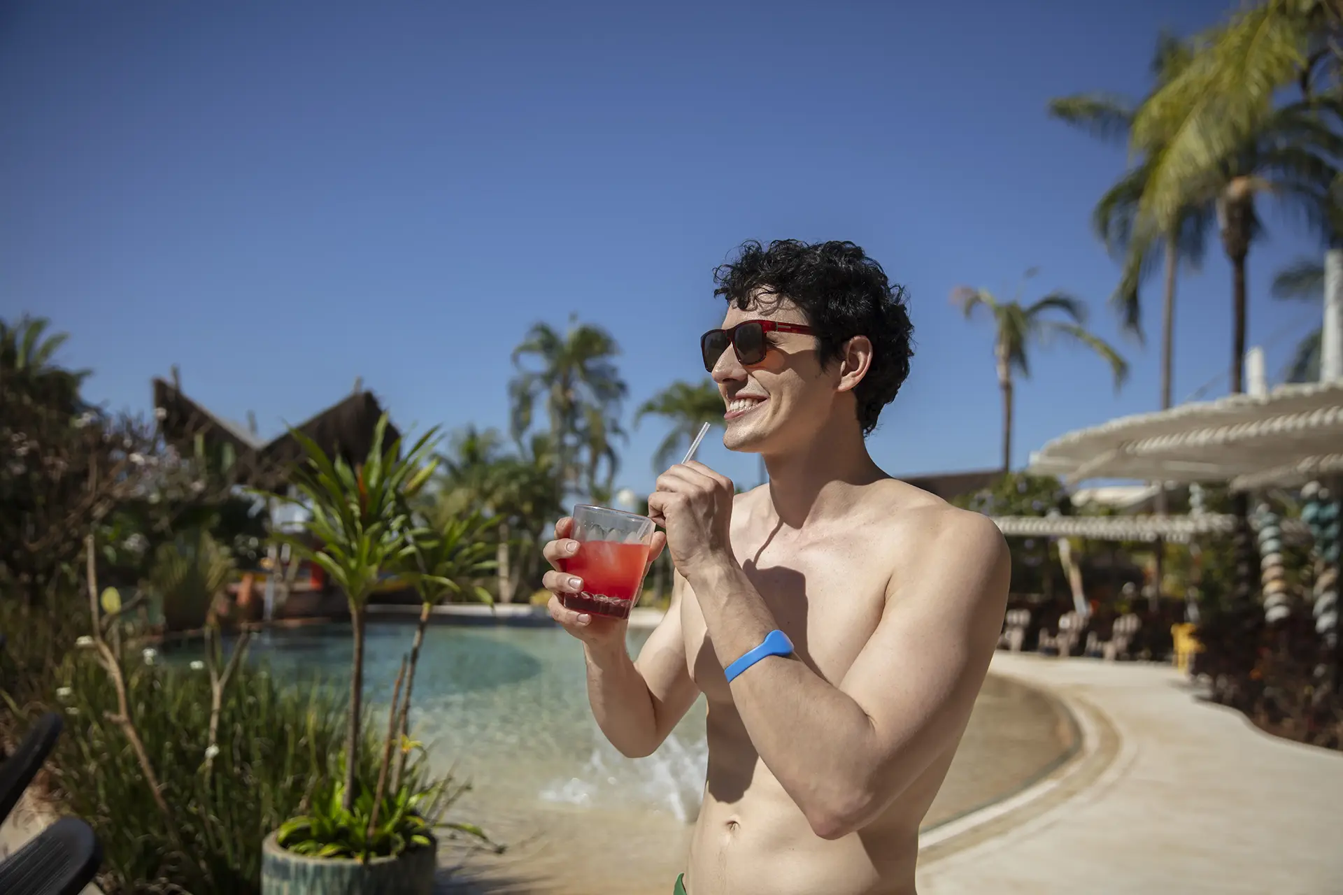Homem relaxando na areia da praia artificial, com coqueiros e quiosques ao fundo.