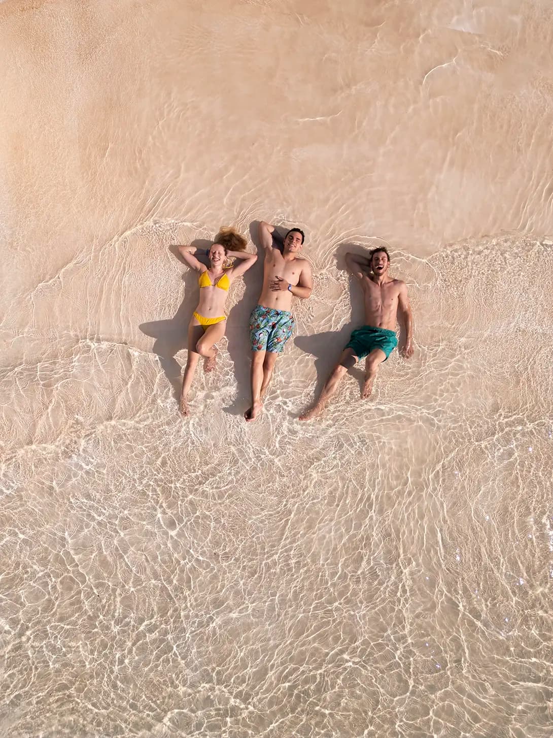 Grupo de amigos deitados na areia, sorrindo para uma foto tirada de cima.