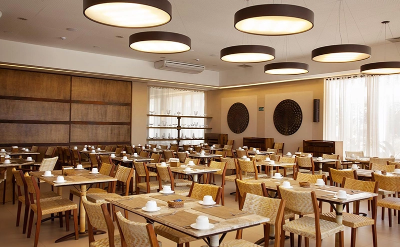Amplo salão do restaurante do hotel com mesas redondas e luminárias circulares.